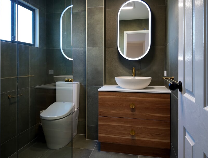 Renovated bathroom with charcoal tiling, a timber vanity and a backlit oval mirror